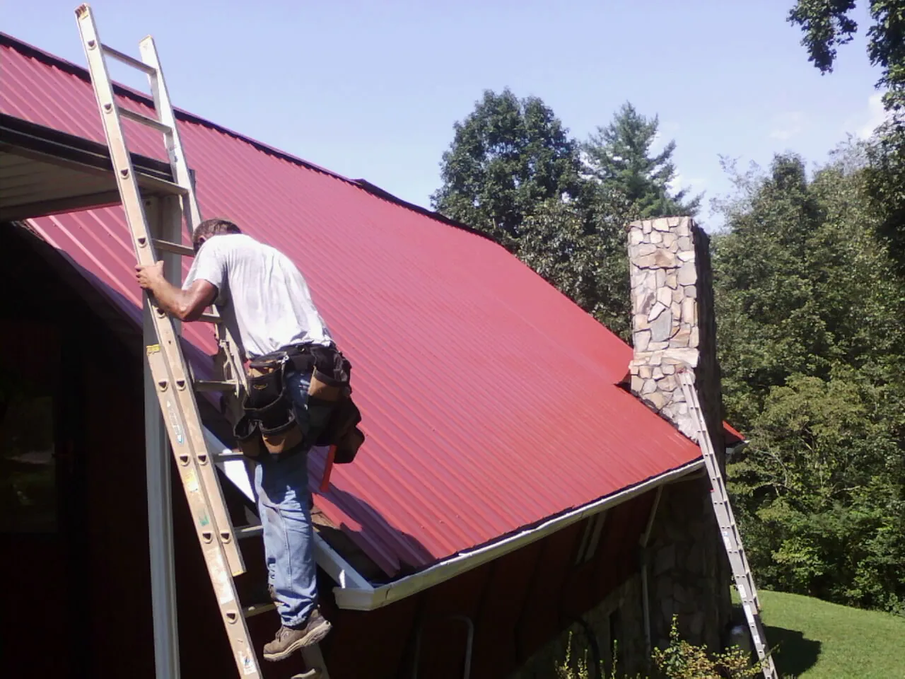 Roofer on a ladder beside a red metal roof and stone chimney — Soffit & Fascia Repair by Roofing Brothers in Callensburg